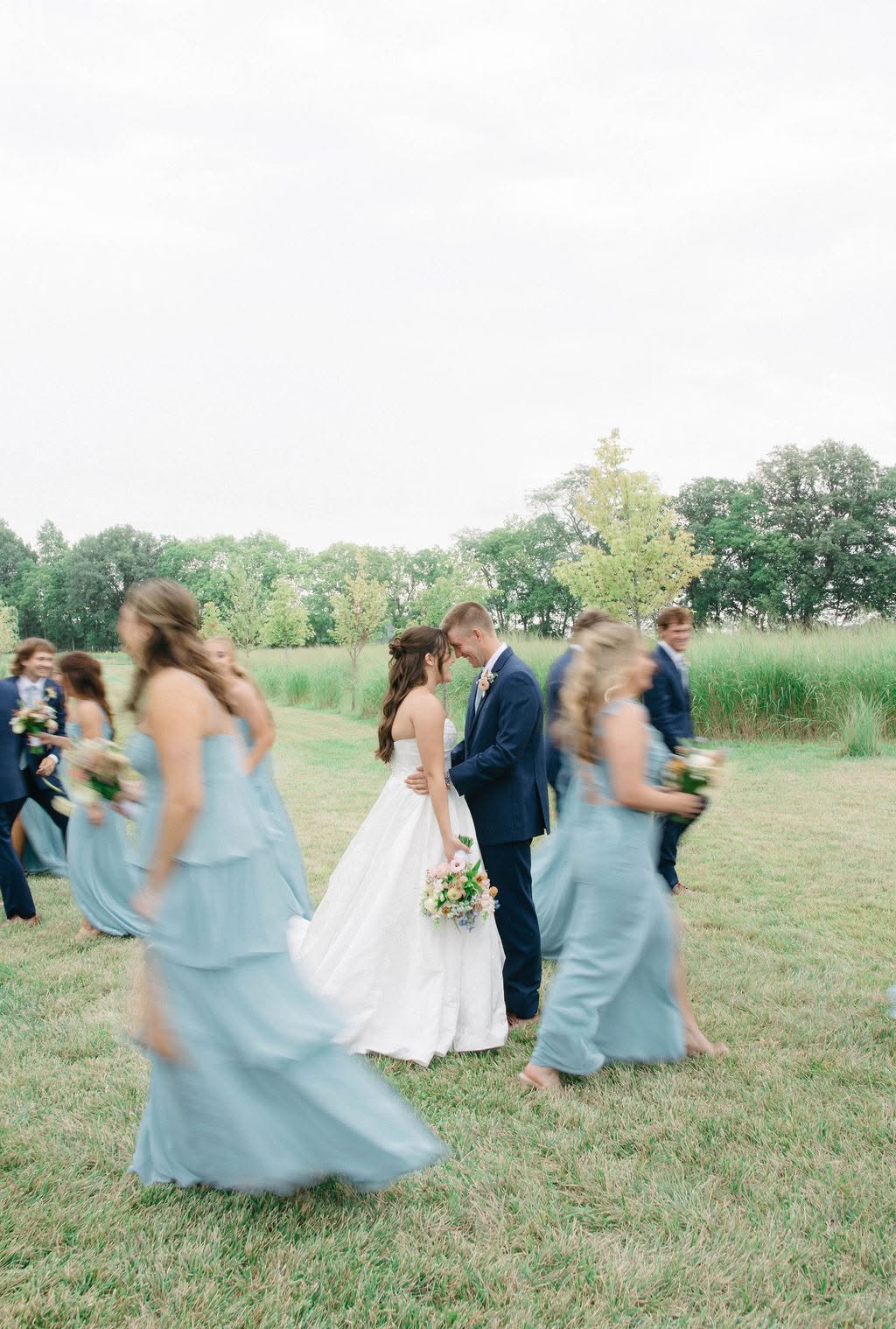 Adisyn and Carson share a moment in the field as their wedding party in dusty blue and navy walks around them
