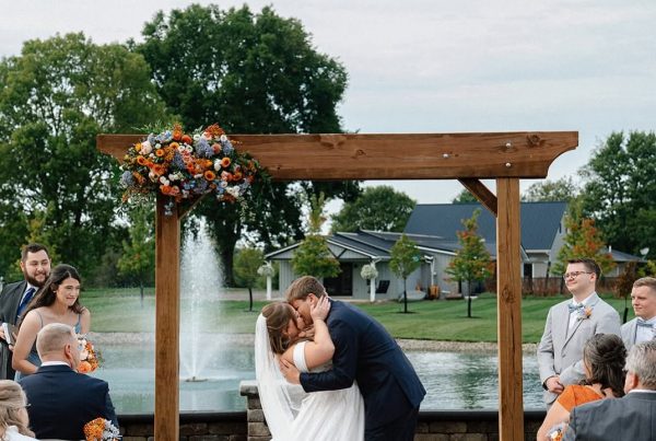 Ashley and Brenton kiss under a wooden arbor with orange and blue florals in front of the pond and fountain