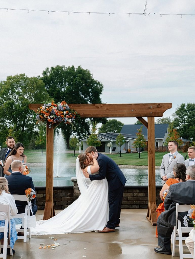 Ashley and Brenton kiss under a wooden arbor with orange and blue florals in front of the pond and fountain