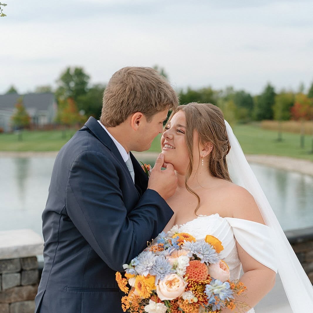 Brenton gently lifts Ashley’s chin by the pond as she holds an orange, peach and dusty blue fall bouquet