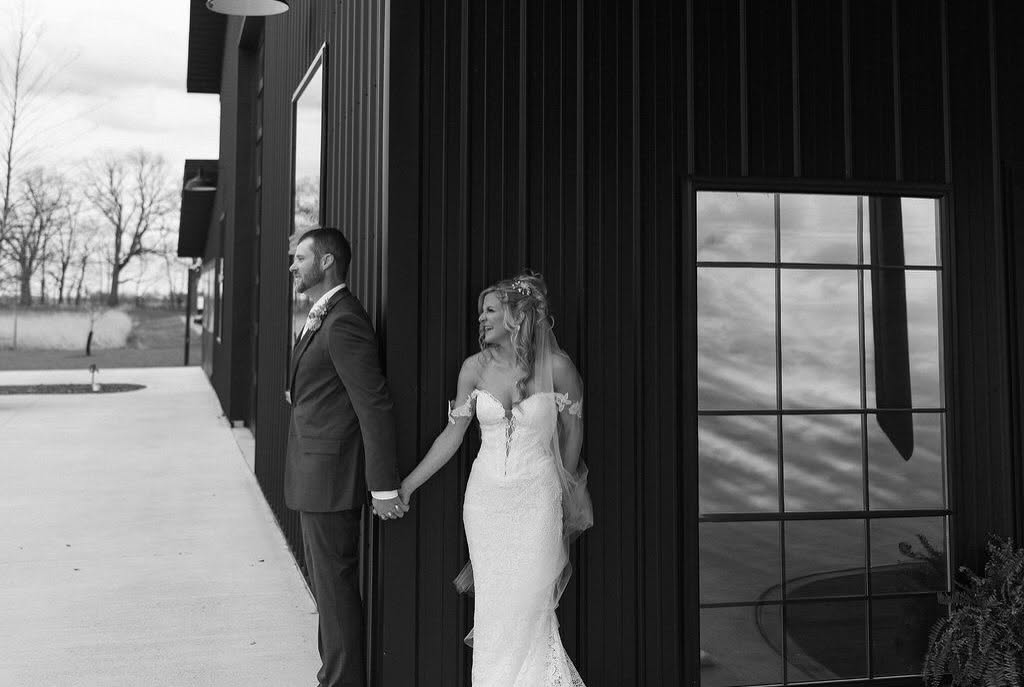 Black and white portrait of Ashley and Chris holding hands beside The Grange Hall barn