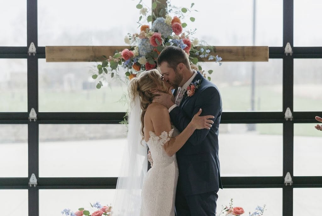 Ashley and Chris share their first kiss in front of a floral wooden cross at The Grange Hall