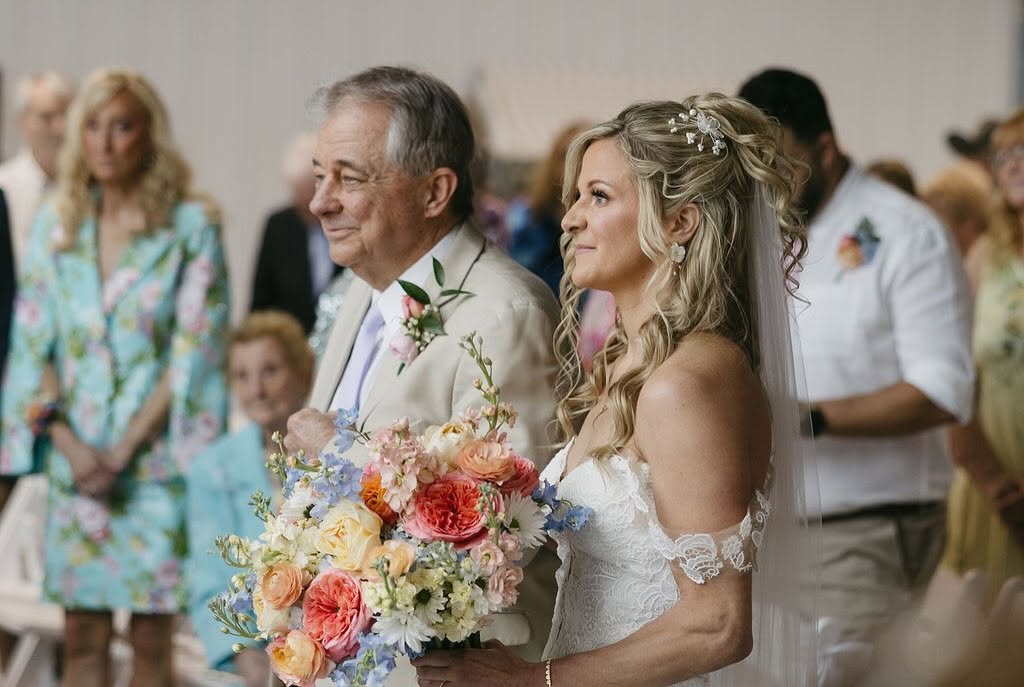 Ashley walks down the aisle with her father carrying a pastel bouquet of peach, pink and blue blooms