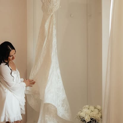 Ava in a white robe admiring her lace wedding gown hanging in the bridal suite