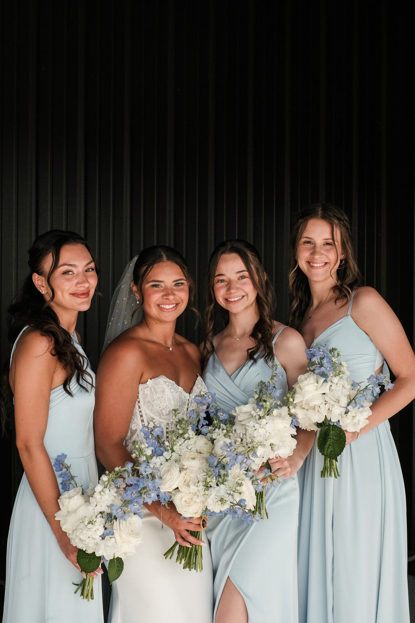 Faith with her bridesmaids in dusty blue dresses holding white rose and blue delphinium bouquets