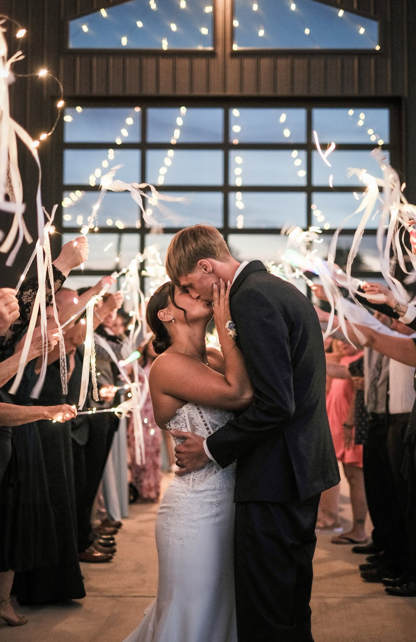 Faith and Nich kiss during their ribbon wand send-off under string lights at The Grange Hall