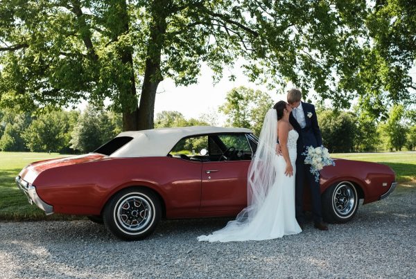 Faith and Nich kiss beside a vintage red convertible under a large shade tree at The Grange Hall