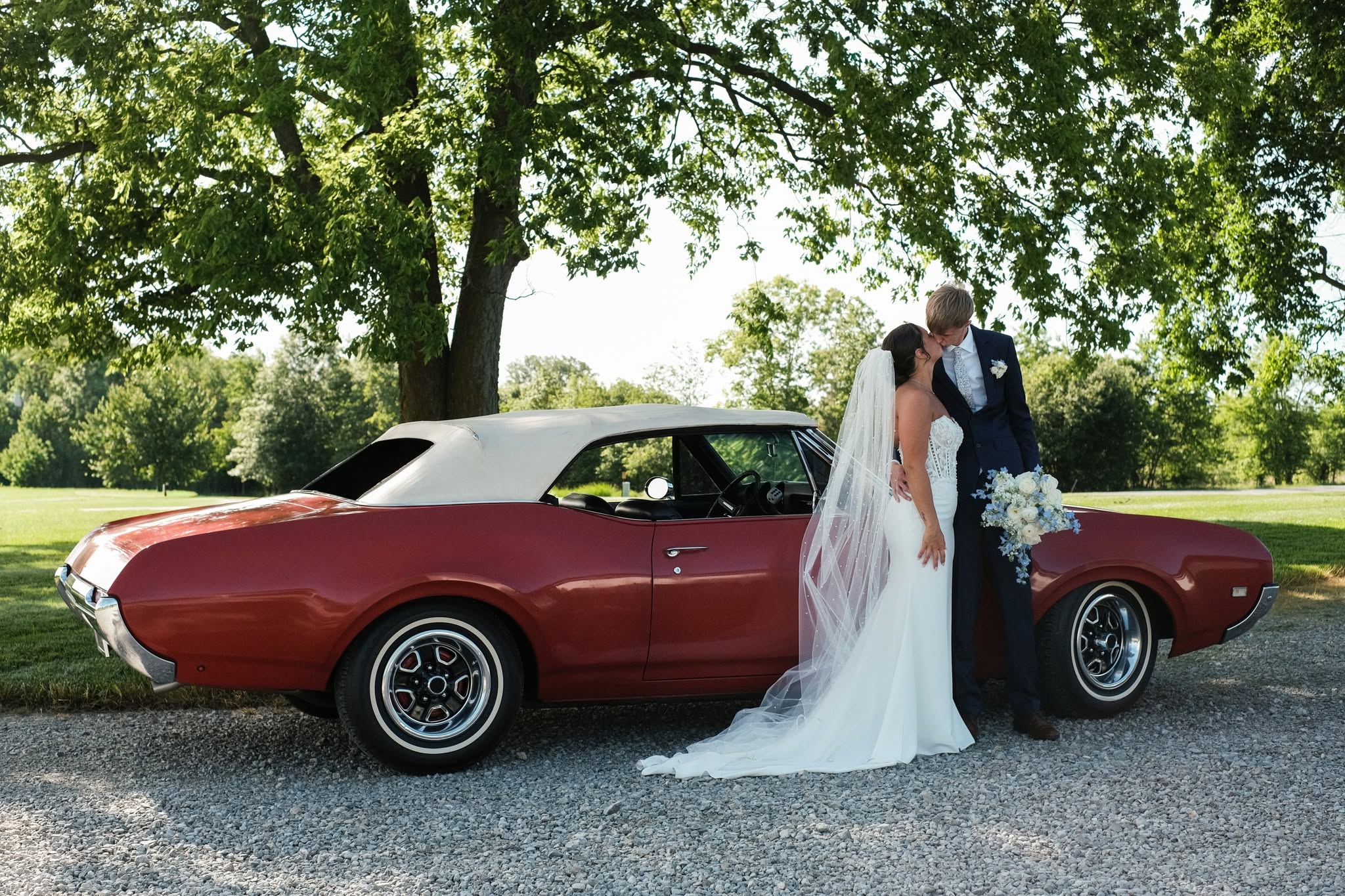 Faith and Nich kiss beside a vintage red convertible under a large shade tree at The Grange Hall
