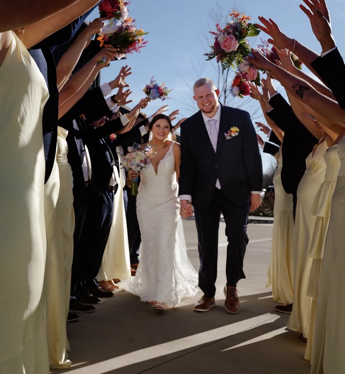 Maddie and Ethan walk through a tunnel of raised bouquets held by their bridal party in butter yellow dresses