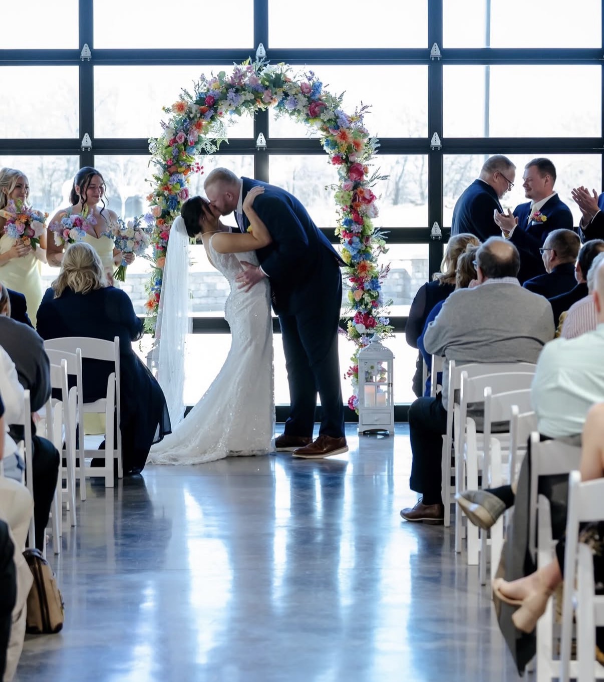 Maddie and Ethan kiss under a colorful floral arch in front of the garage door windows during their indoor ceremony