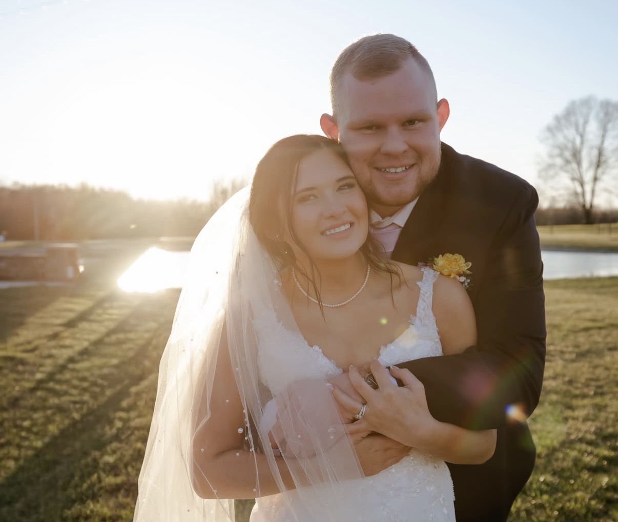 Maddie in a pearl necklace and veil embraced by Ethan in a golden hour portrait by the pond