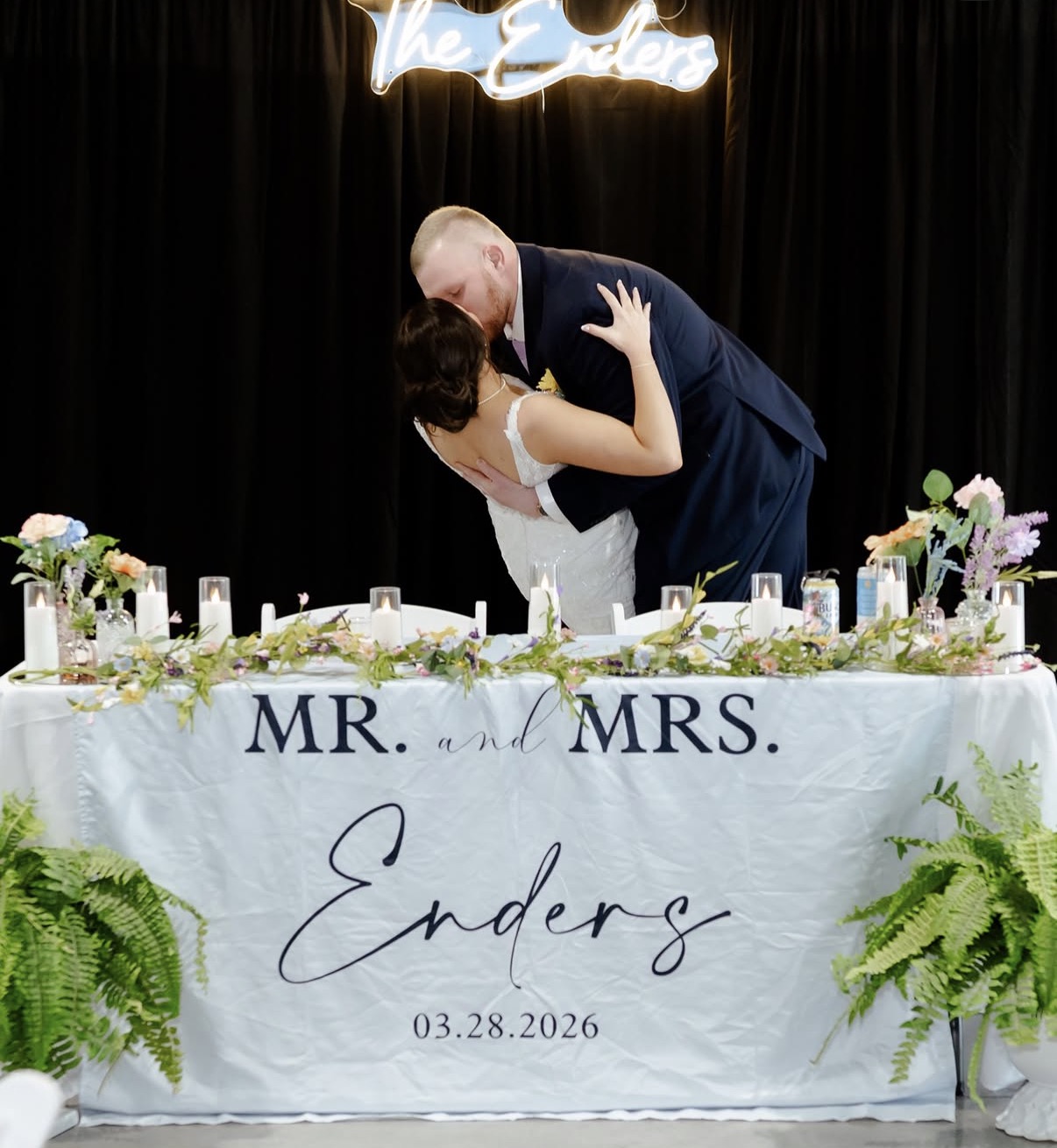 Maddie and Ethan kiss behind their sweetheart table with a floral garland and custom neon sign
