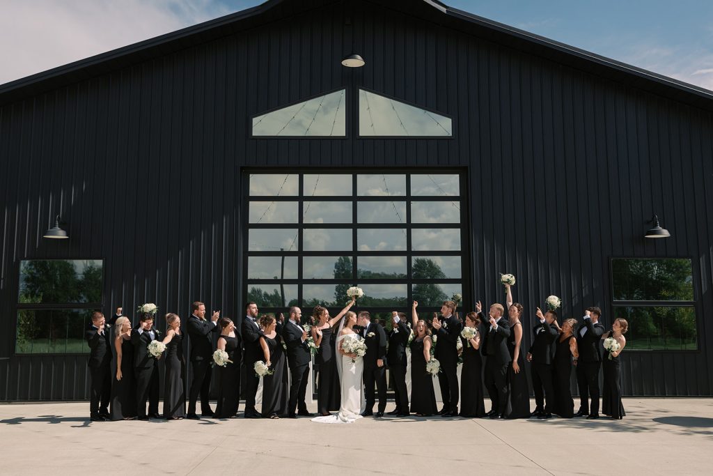 Taylor and Derek kiss as their large wedding party in black cheers in front of The Grange Hall barn