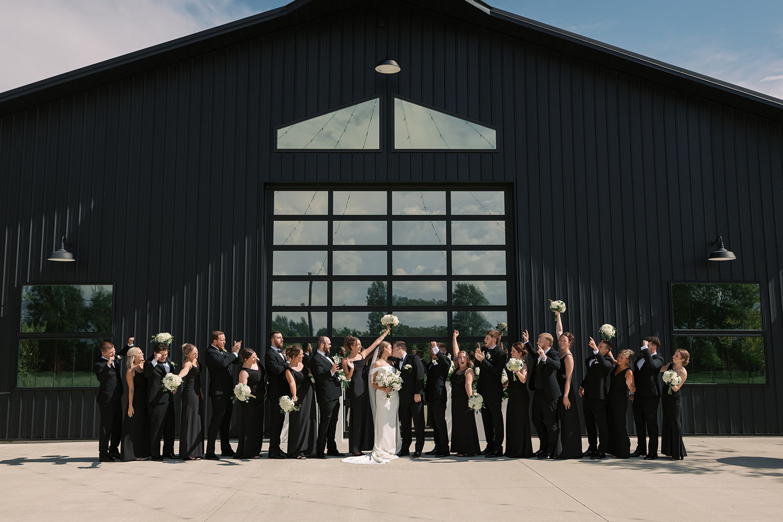 Taylor and Derek kiss as their large wedding party in black cheers in front of The Grange Hall barn